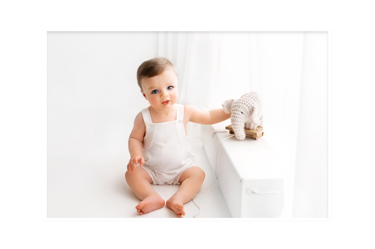 baby boy on white backdrop in white romper playing with knitted elephant toy